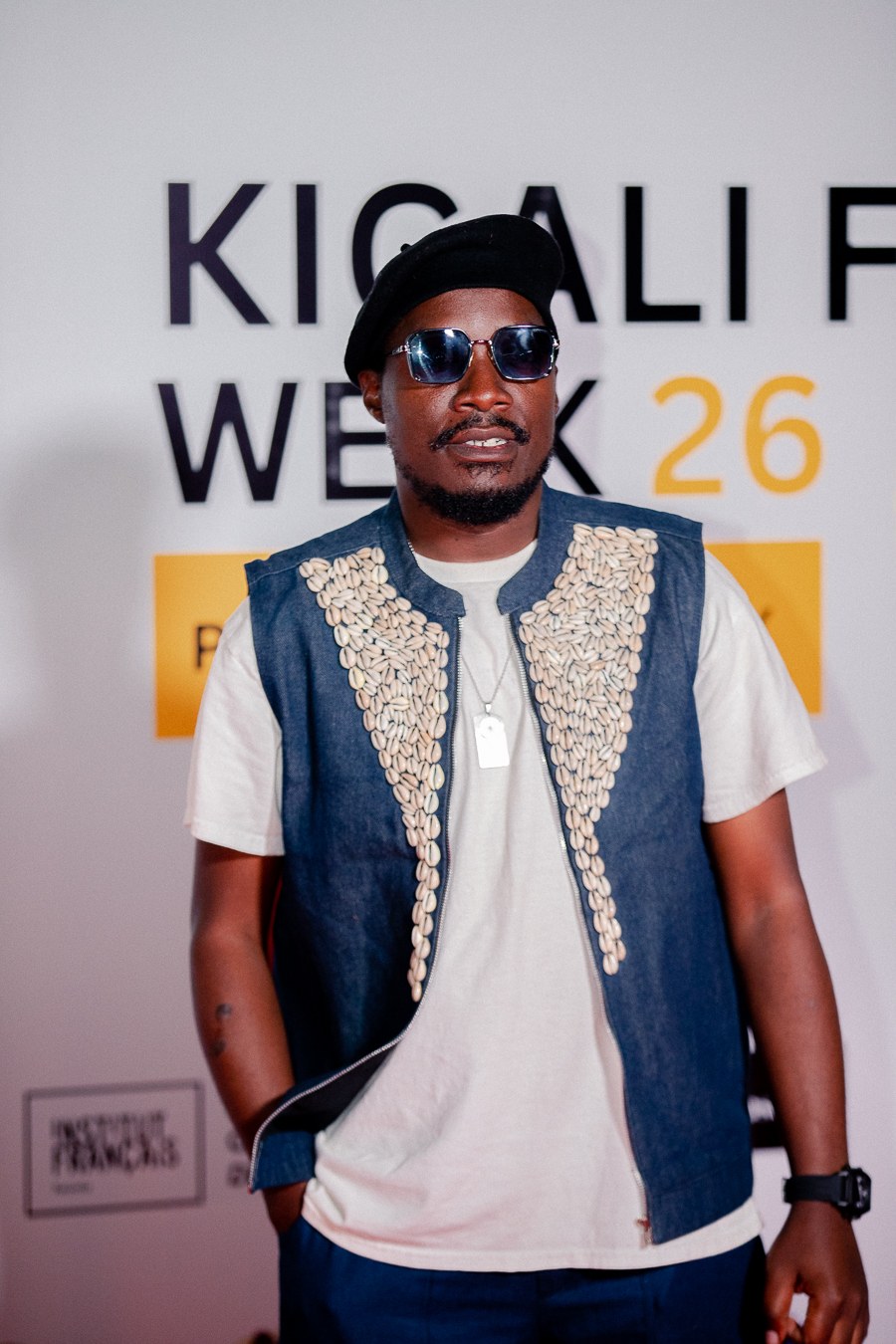 Mukasa Najib — The Fabric Murderer — at Kigali Fashion Week 2026 step and repeat, wearing cowrie shell denim vest and beret